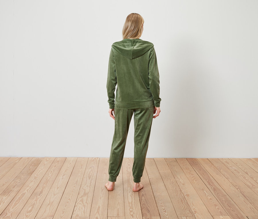Bir kadın kameraya arkası dönük duruyor ve zeytin yeşili renginde kadife bir eşofman takımı giyiyor.