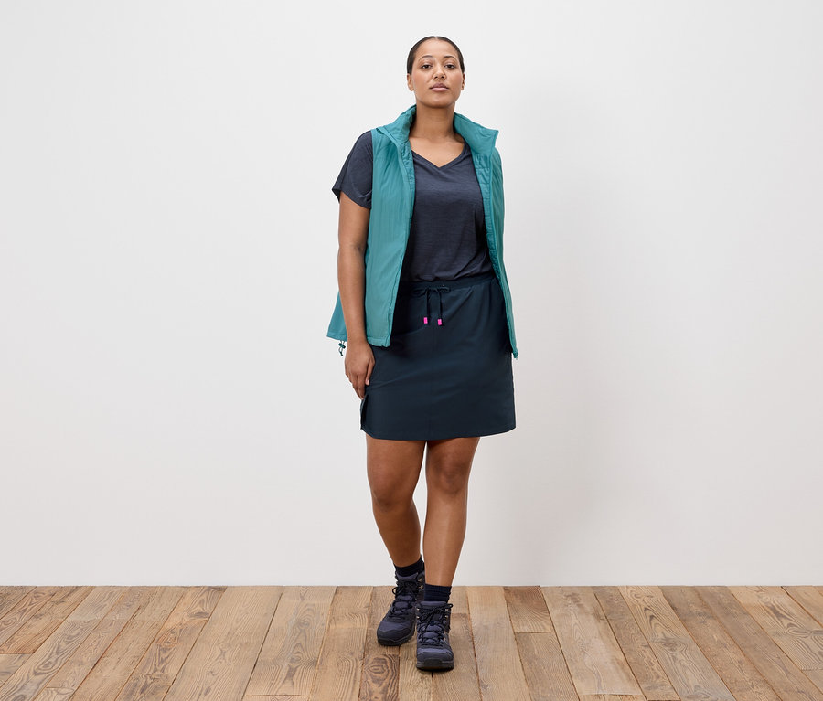 Kadın beyaz bir duvarın önünde duruyor, lacivert bir etek, lacivert bir tişört, petrol mavisi bir yelek ve lacivert yürüyüş ayakkabıları giyiyor.