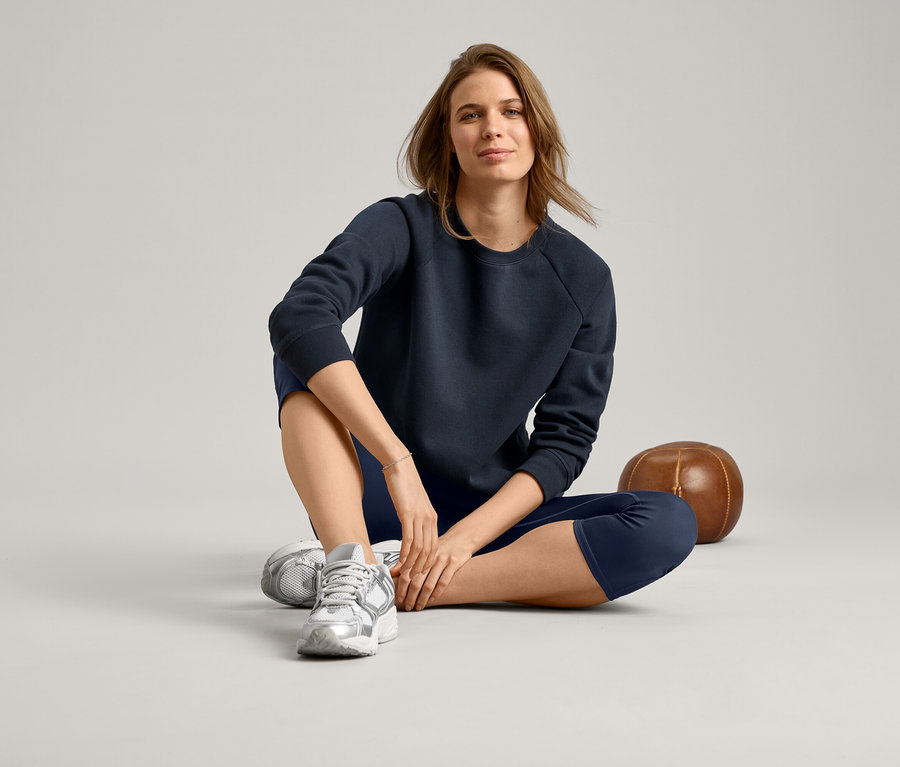 Kadın bağdaş kurmuş oturuyor, lacivert bir sweatshirt, lacivert tayt ve gümüş spor ayakkabısı giyiyor.