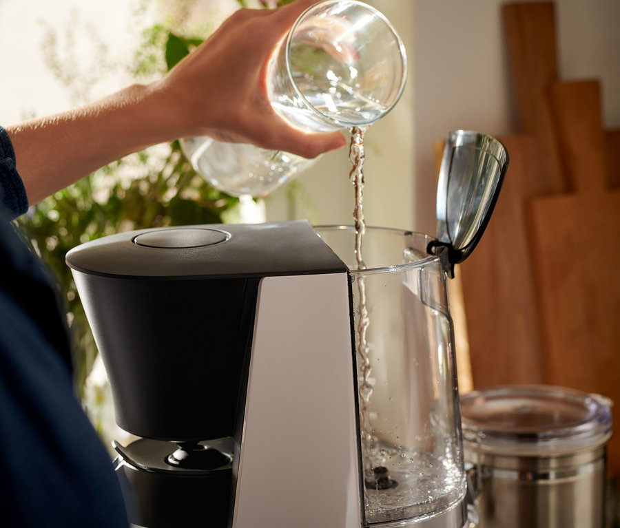 Kadın, şeffaf bir bardaktan gri-siyah »Let's Brew« Filtre Kahve Makinesi'ne su döküyor.