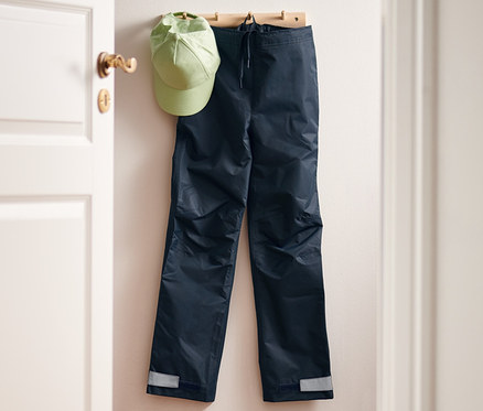 Askıda bir yağmur pantolonu ve yeşil bir şapka asılı duruyor. Paspasın üzerinde bir yağmur pantolonu ve yanında lastik çizme duruyor.