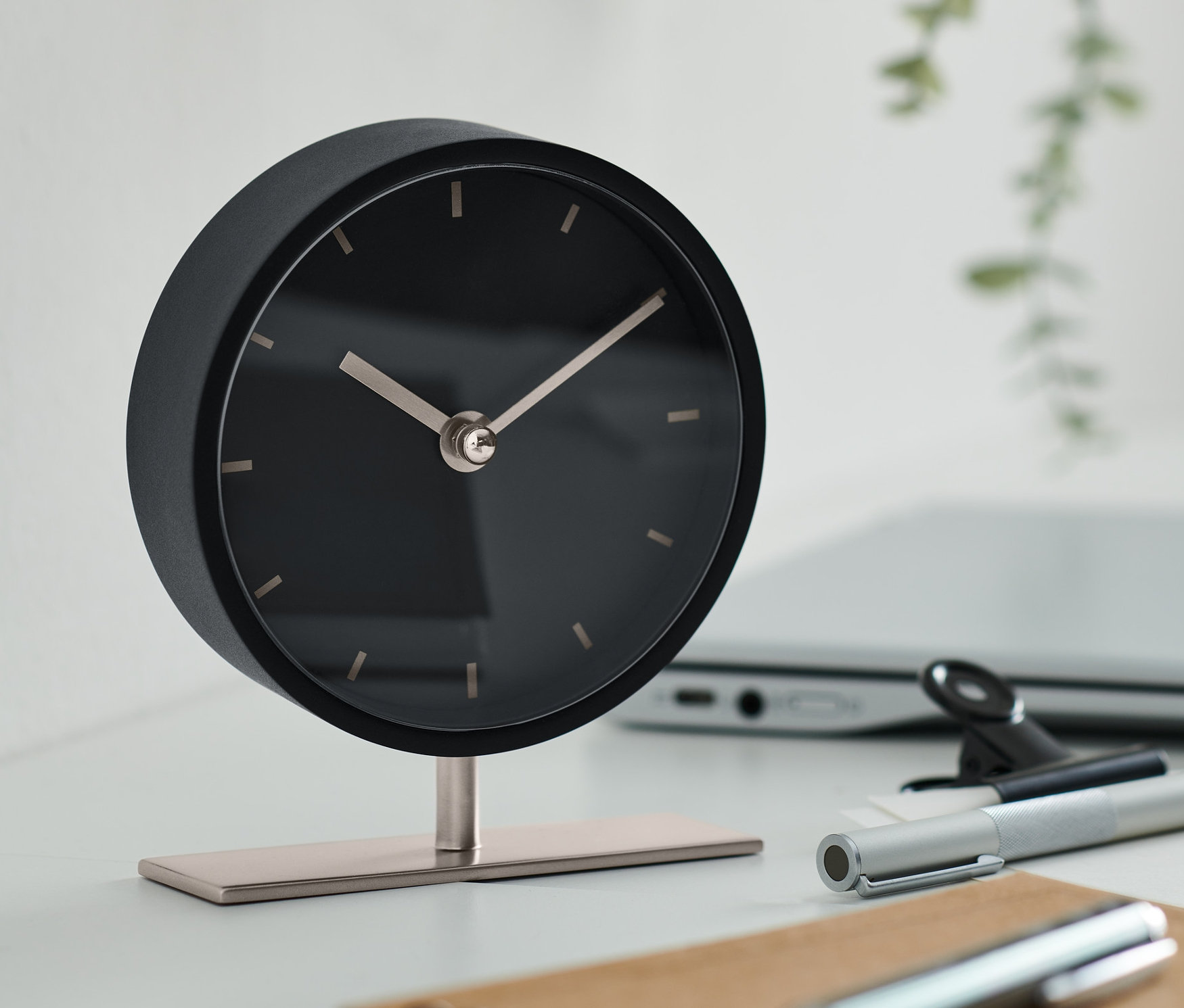 Siyah tasarım masa saati, dizüstü bilgisayar, kalem ve not defteri olan bir masanın üzerinde duruyor.