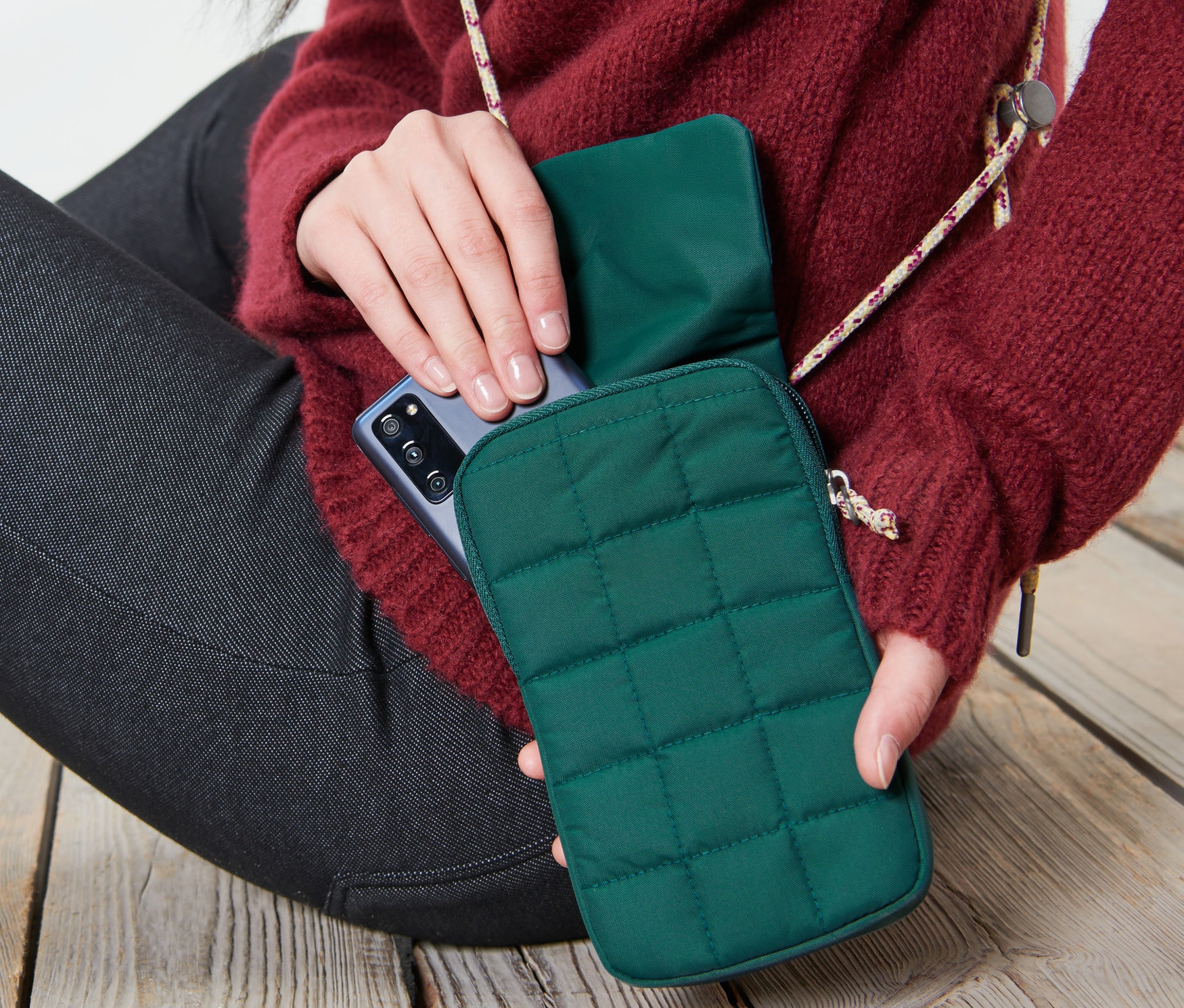 Kadın ahşap zeminde oturuyor ve bir telefonu koyu yeşil yalıtımlı akıllı telefon çantasına koyuyor.