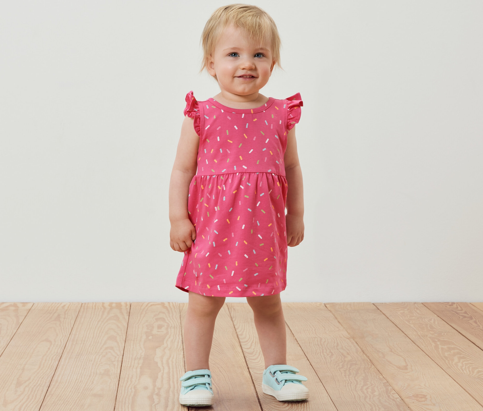 Gülümseyen küçük bir kız pembe bir jarse elbise giyiyor.