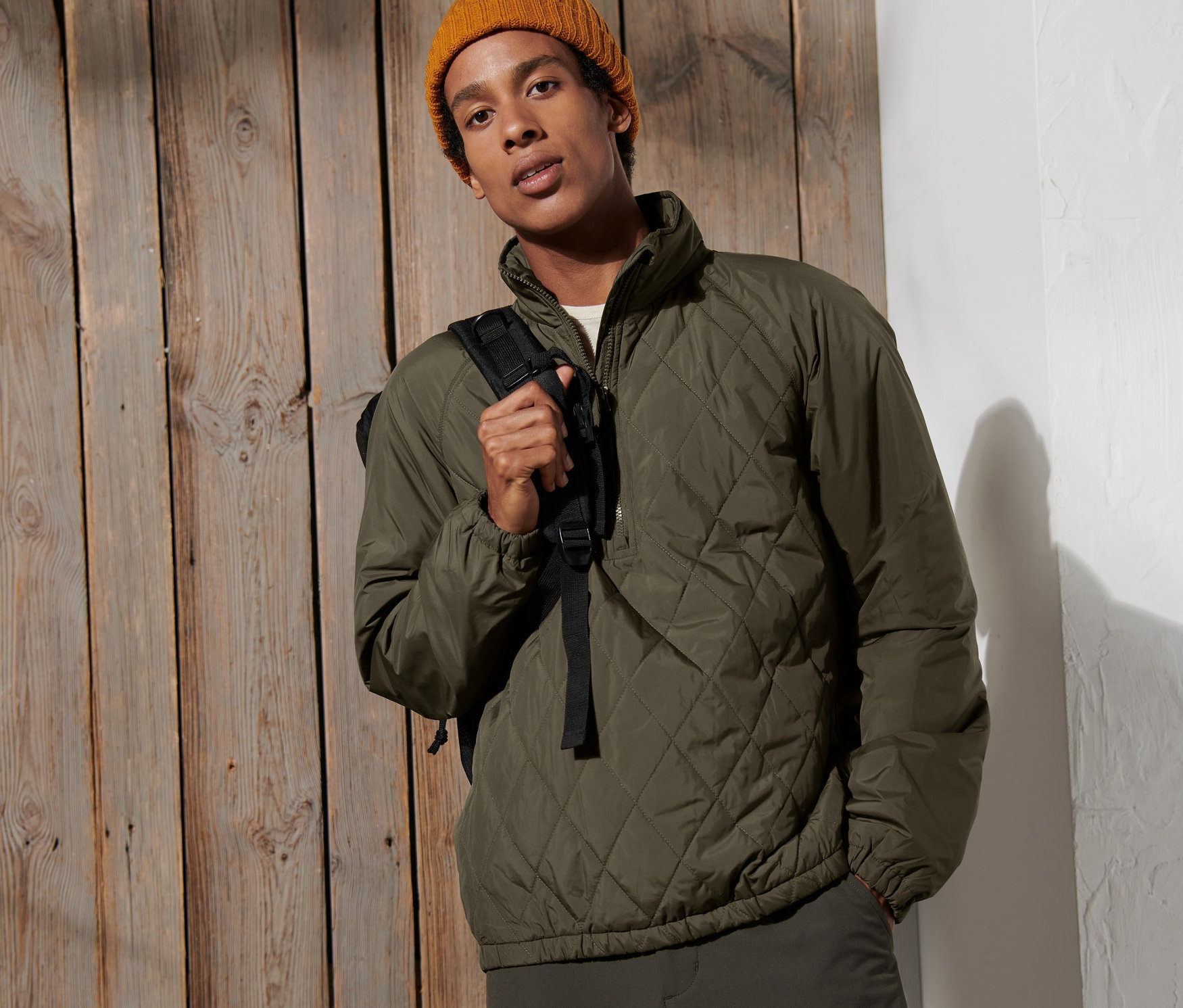 Adam ahşap bir duvarın önünde poz veriyor, Rüzgar Geçirmez Ceket ve sarı bir bere takıyor.