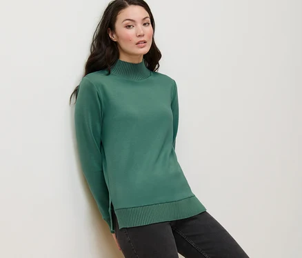 Bir kadın duvara yaslanmış, üzerinde yeşil yarım balıkçı yaka sweatshirt var.
