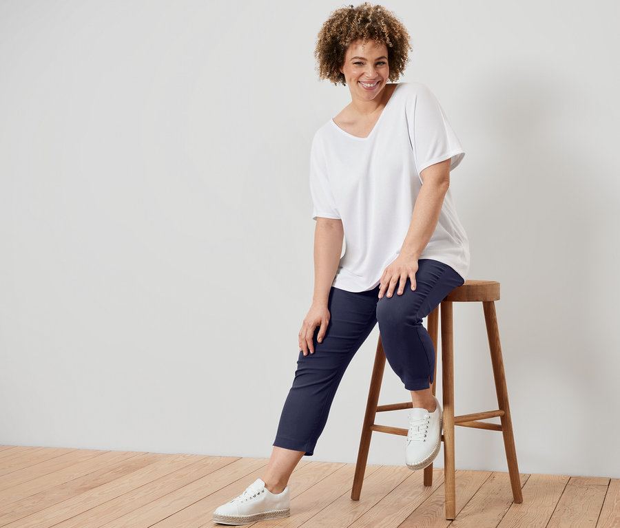 Bir kadın taburede oturuyor, beyaz dokulu tişört ve lacivert 7/8 streç pantolon giyiyor.