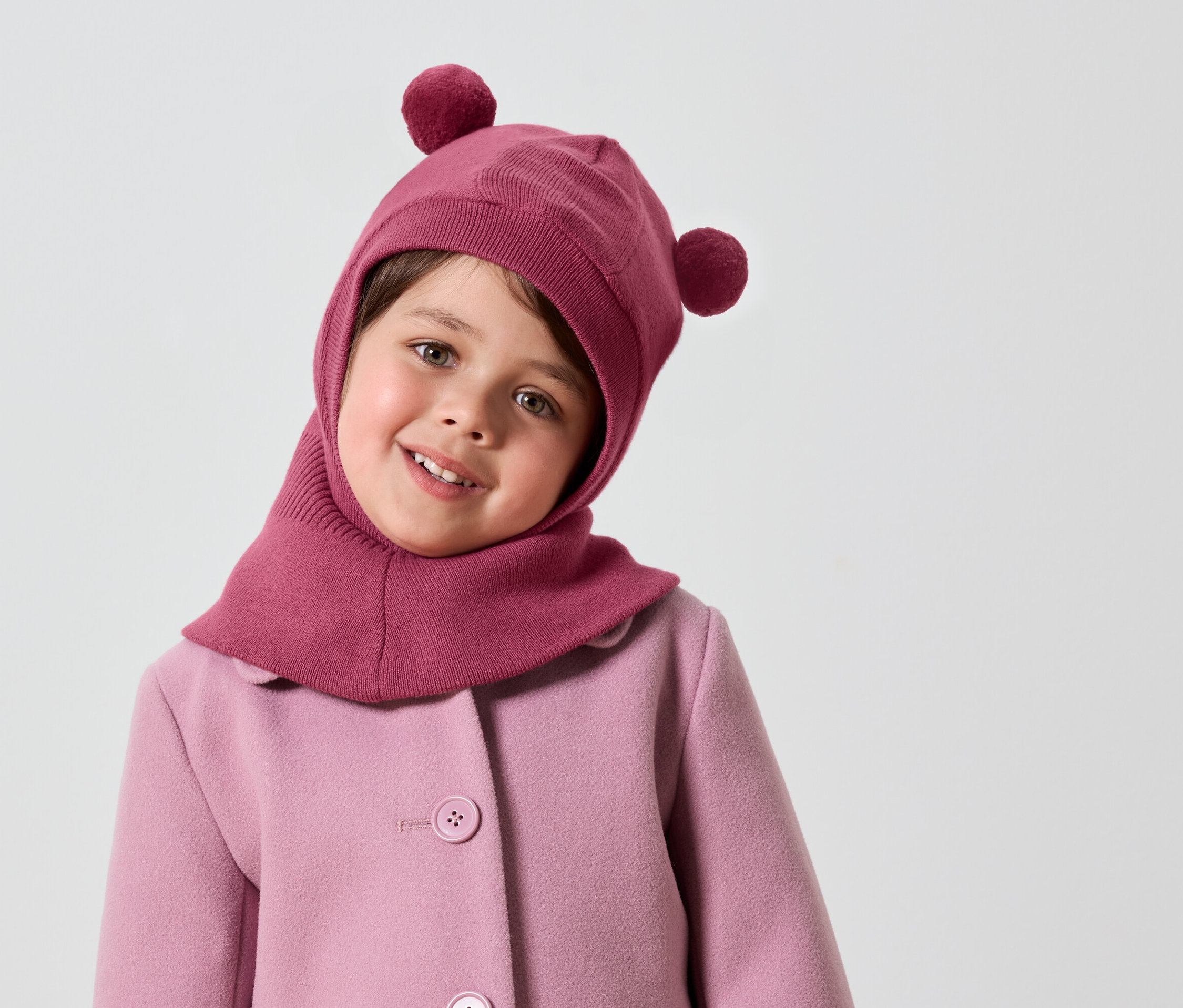 Ponponlu pembe başlığı ve pembe mantosuyla gülümseyen küçük kız çocuğu.