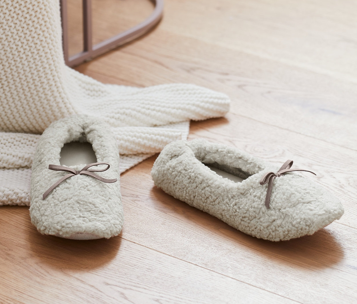 Ahşap zeminde, beyaz örgü battaniye ve pembe taburenin yanında krem rengi bir çift Ev Ayakkabısı bulunmaktadır.