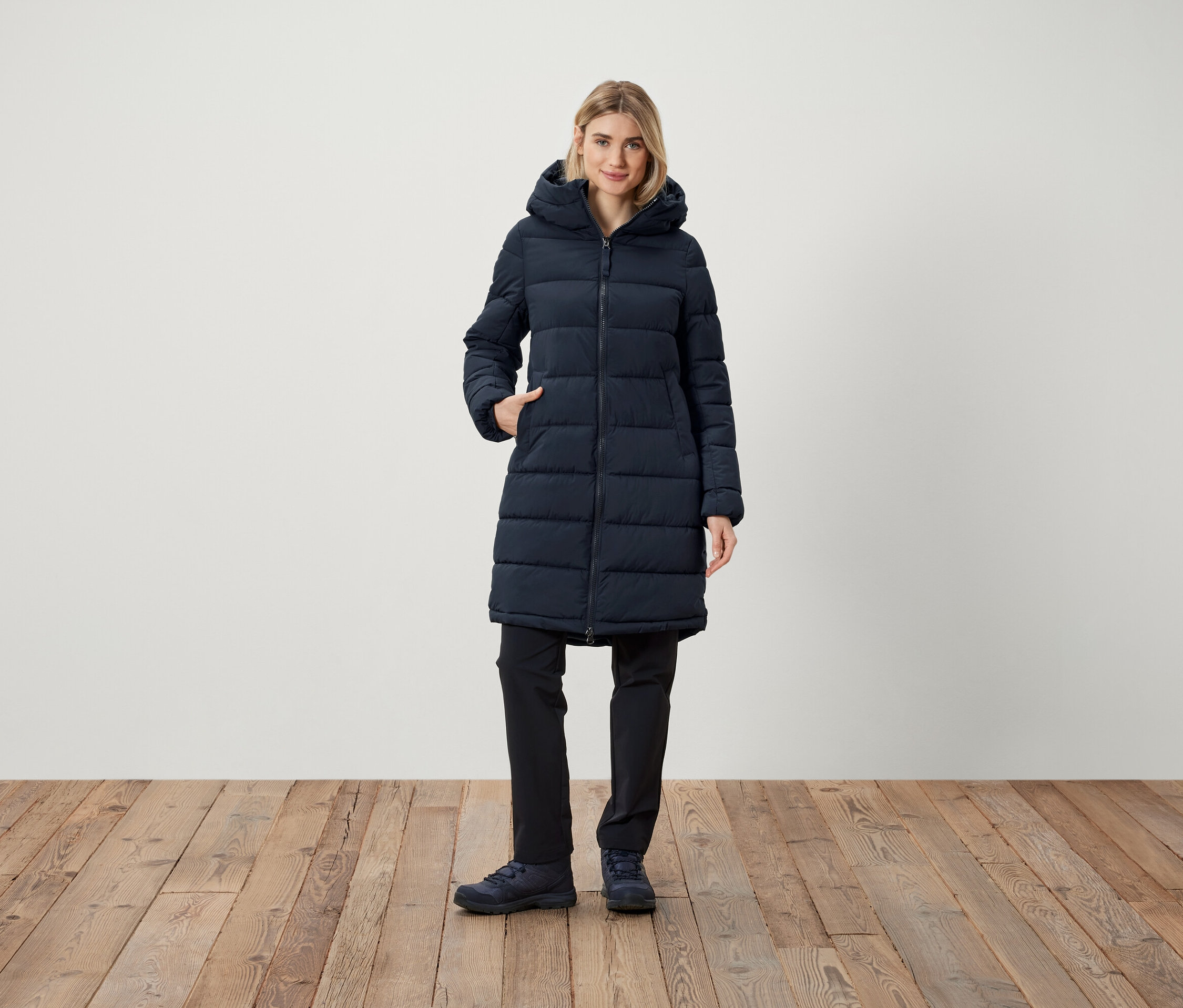 Kadın, kapüşonlu, lacivert, şişme montunun cebinde eliyle duruyor.