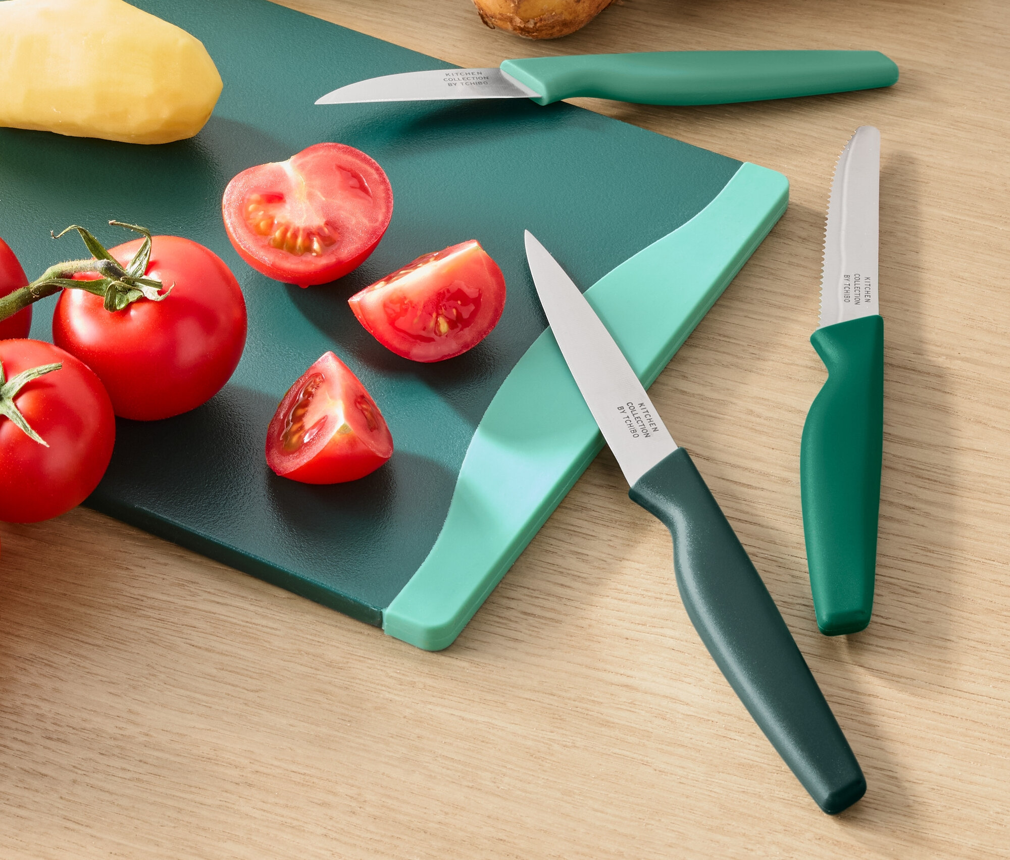 Ahşap bir masanın üzerinde domateslerin bulunduğu yeşil bir kesme tahtası ve koyu yeşil saplı üç bıçak bulunmaktadır. Tahtanın etrafında patatesler, sarı bir biber ve kabak bulunmaktadır.