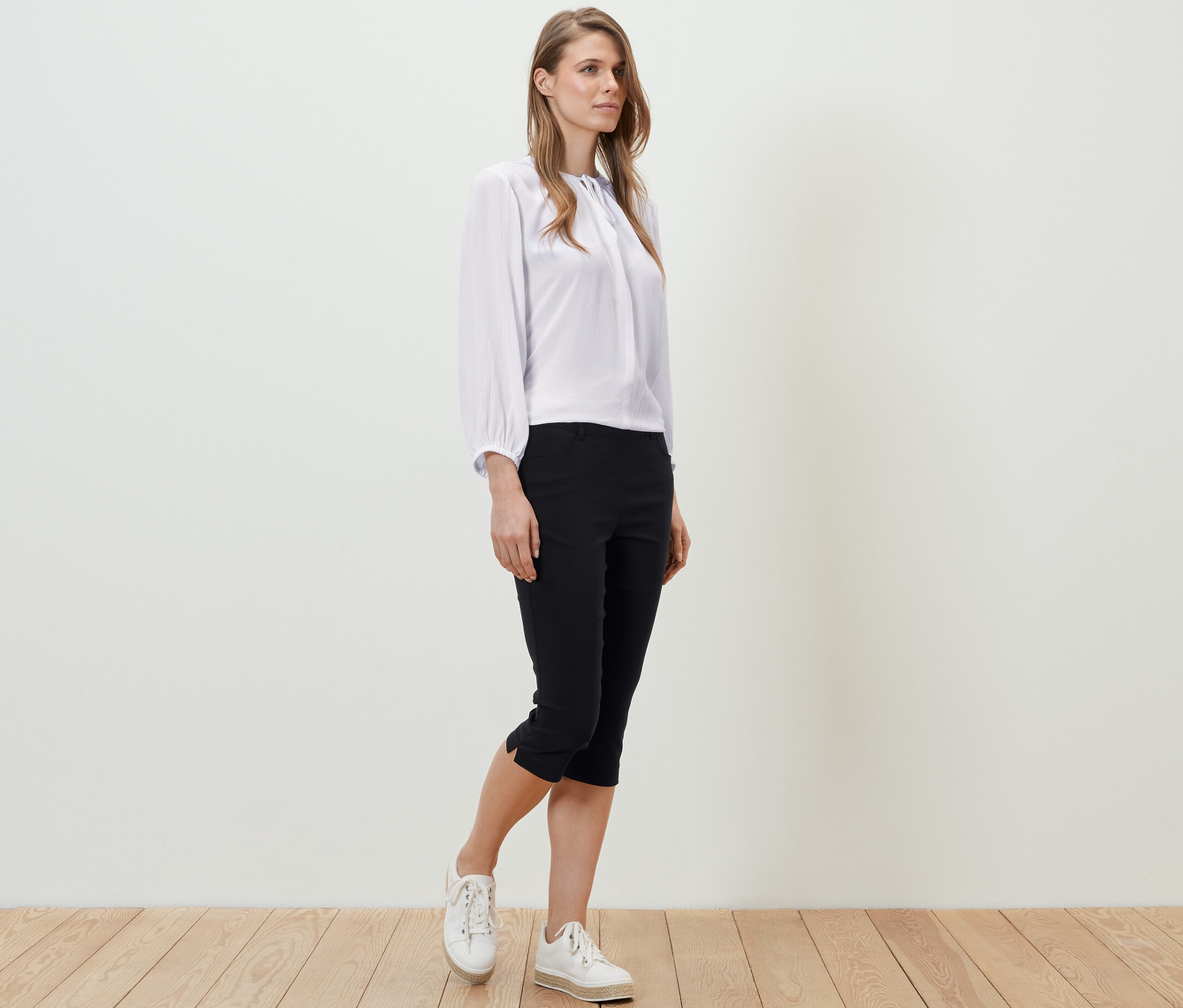 Ahşap zeminde duran bir kadın, beyaz bir bluz ve siyah 3/4 Bengalin Pantolon giyiyor.