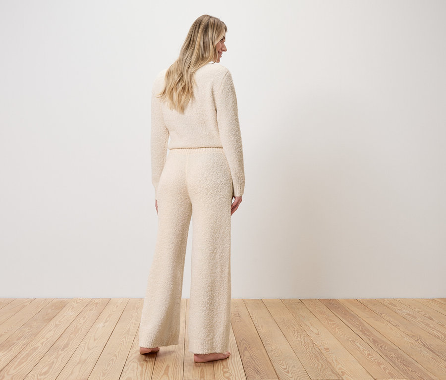 Kadın arkası dönük duruyor, buklet örgülü kazak ve geniş paçalı pantolon giyiyor.