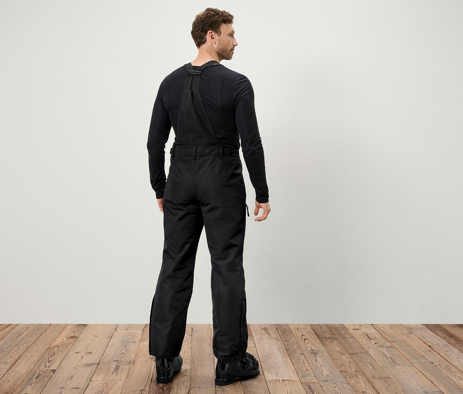 Arkası dönük bir adam, askılı siyah kayak pantolonu ve siyah kayak botları giyiyor.