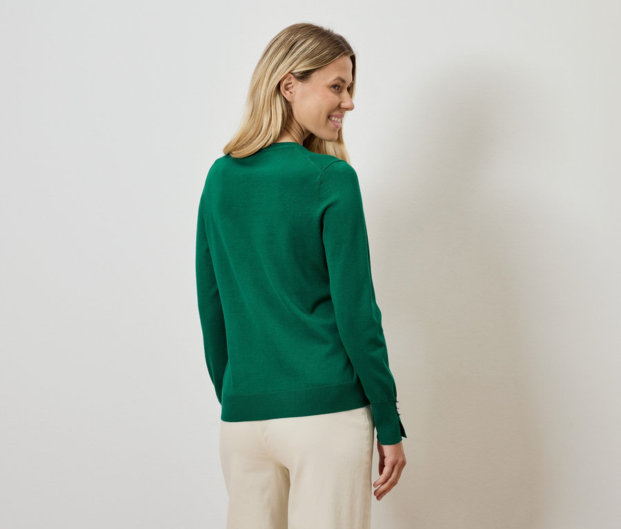 Kadın omzunun üzerinden bakıyor, üzerinde yeşil bir kazak ve bej pantolon var.