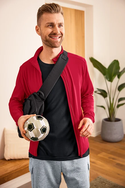 Kırmızı Funktionsjacke, siyah Sportshirt, Cargo-Sweatshorts, Crossbody-Bag ve elinde Fußball olan adam.