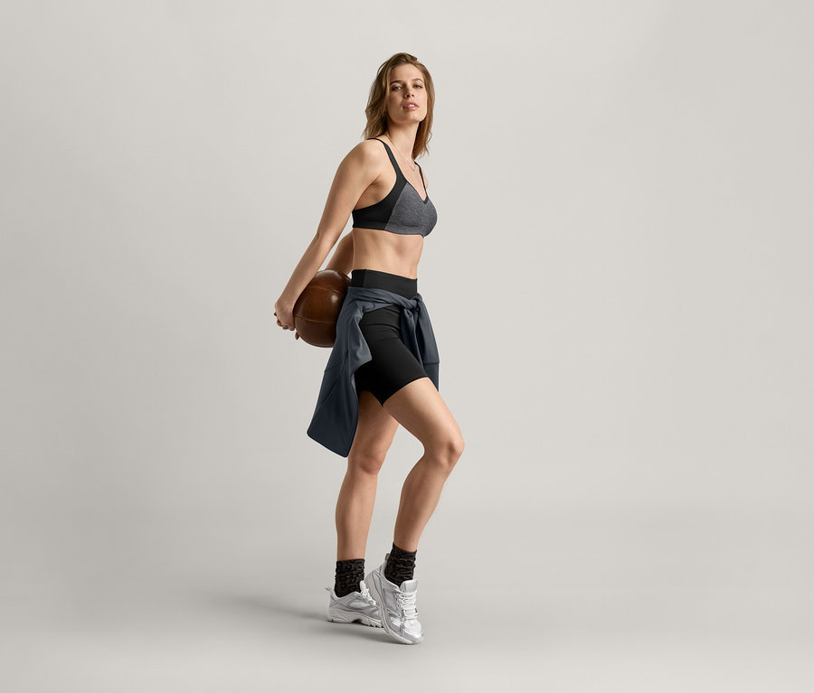 Kadın sırtında basketbol topu tutuyor, spor sütyeni, şort ve antrasit spor ceket giyiyor.