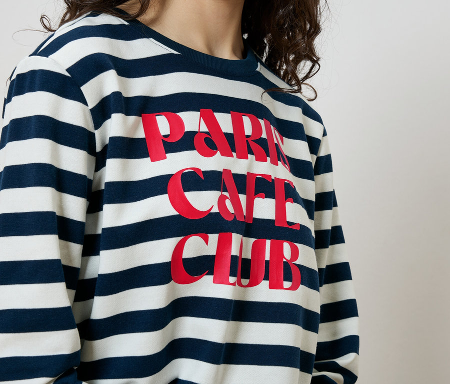 Kadın, üzerinde kırmızı renkte "Paris Cafe Club" yazısı olan mavi-beyaz çizgili bir sweatshirt giyiyor.