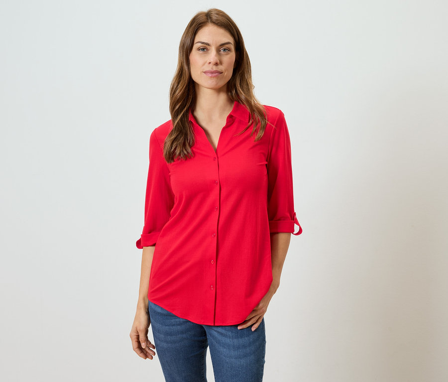 Kahverengi saçlı bir kadın kırmızı bir bluz ve mavi kot pantolon giyiyor.