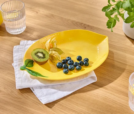 Sarı tabakta yaban mersini, kivi ve altın çilek beyaz bir bez üzerinde duruyor. Yanında limonlu bir bardak su ve bir limon var.
