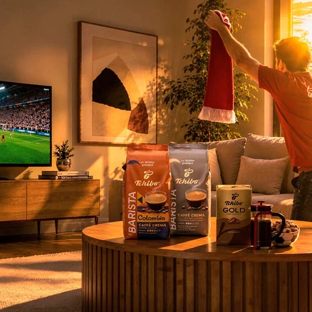 Kırmızı tişörtlü bir adam, televizyonda futbol maçı izlerken kırmızı-beyaz bir atkı kaldırıyor. Sehpanın üzerinde iki Tchibo Barista kahve paketi, bir Tchibo Gold kutusu, bir French Press ve bir kase kuruyemiş var.