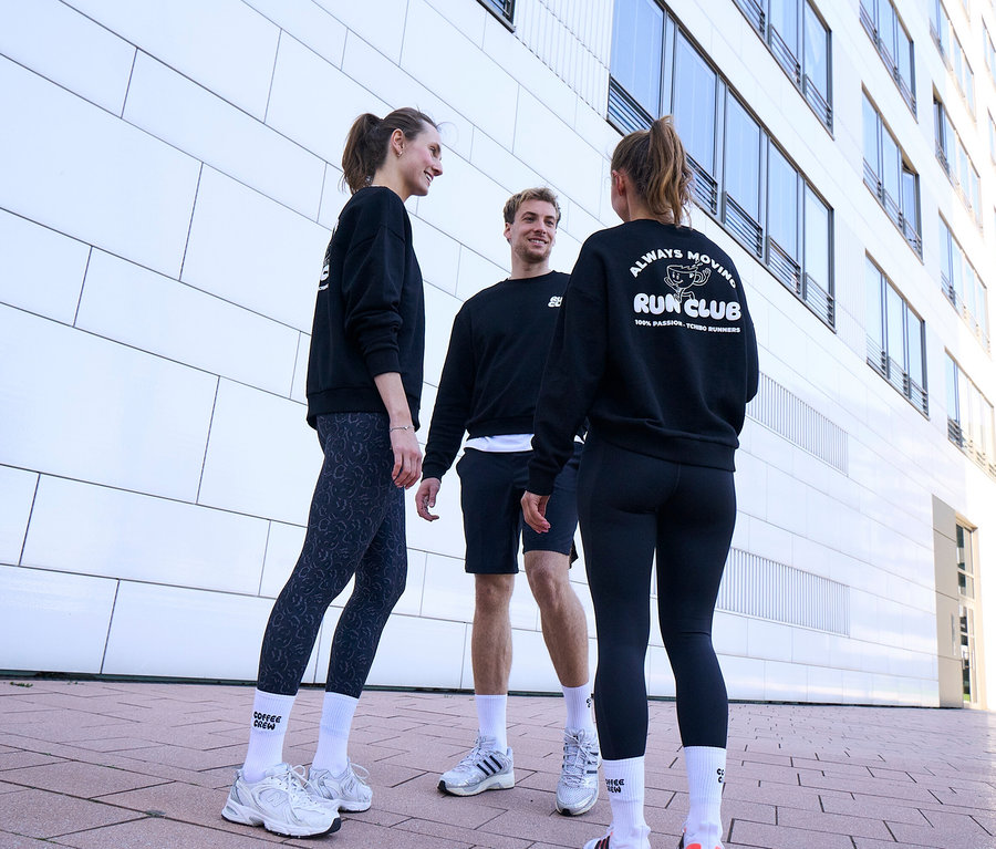 İki kadın ve bir erkek olmak üzere üç kişi gülümsüyor ve sohbet ediyor. Hepsi siyah sweatshirt ve beyaz Unisex Coffee Crew Tenis Çorapları giyiyor. Erkek siyah spor şortu giyiyor. Bir kadın gri-siyah Spor Tayt "ActivePower", diğer kadın ise siyah Spor Tayt "ActivePower" giyiyor.