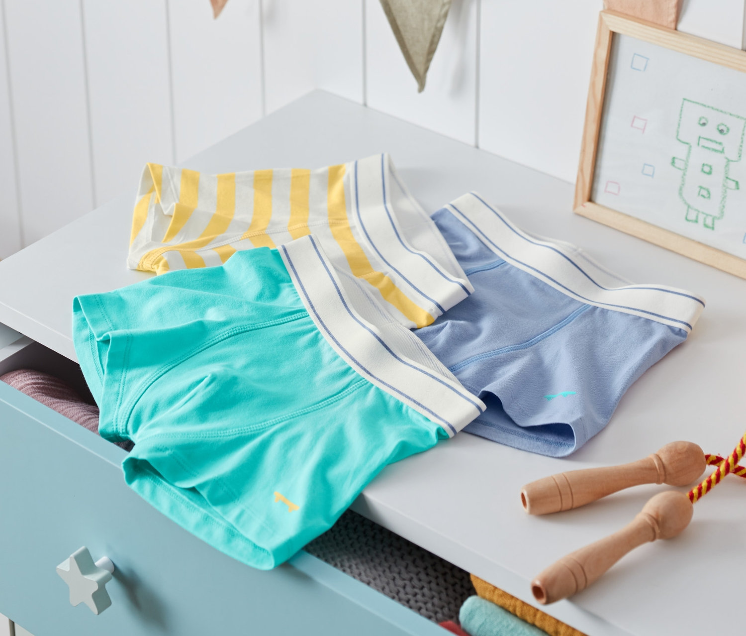 Üç adet organik pamuklu erkek çocuk slip boxer, bir tilki oyuncağı ve bir robot resminin yanında bir şifonyerin üzerinde duruyor.