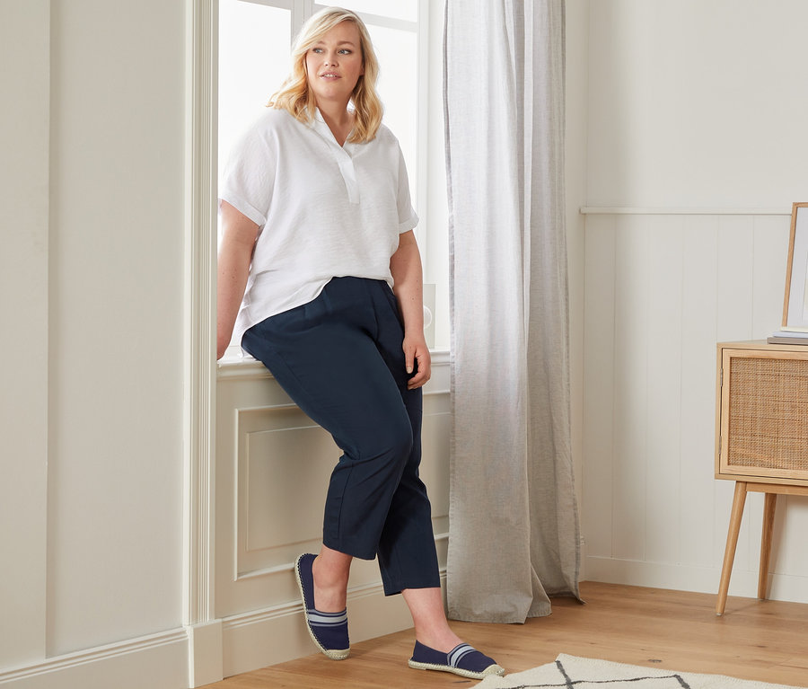 Kadın pencere pervazında oturuyor, üzerinde organik pamuklu oversized bluz, rahat chino pantolon ve espadriller var.