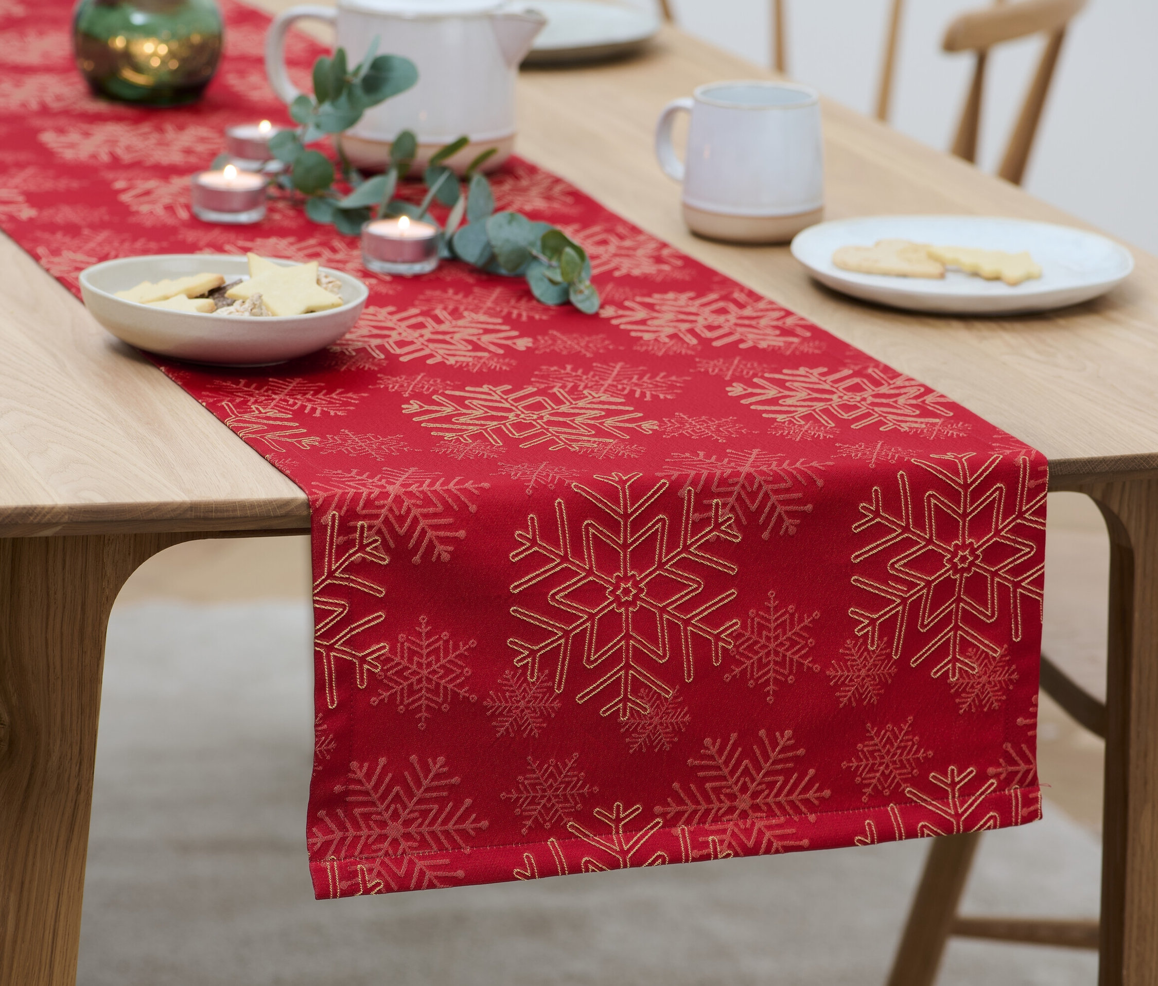 Ahşap bir masa üzerinde kar tanesi desenli kırmızı jakar runner, üzerinde kurabiye tabakları, mumlar ve bir çaydanlık bulunmaktadır.