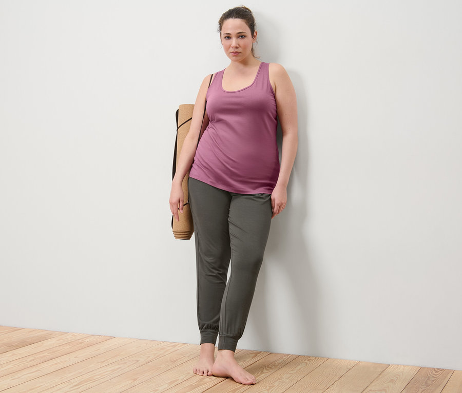Duvara yaslanmış kadın, eflatun 2 adet atlet ve yoga pantolonu giyiyor, omzunda yoga matı var.