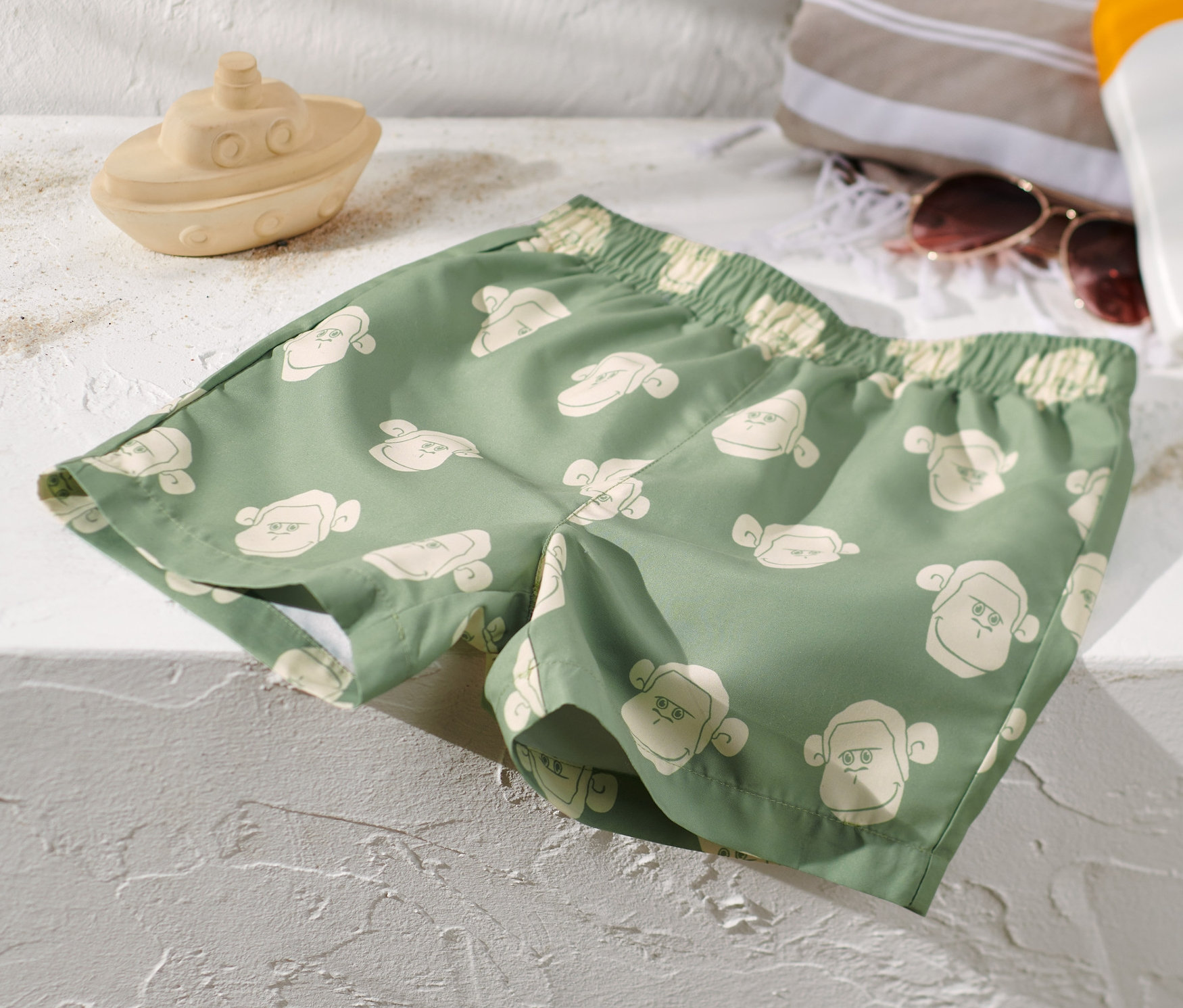 Yeşil, maymun desenli çocuk deniz şortu, oyuncak bir tekne, havlu, güneş gözlüğü ve güneş kremi yanında beyaz bir yüzeyde duruyor.