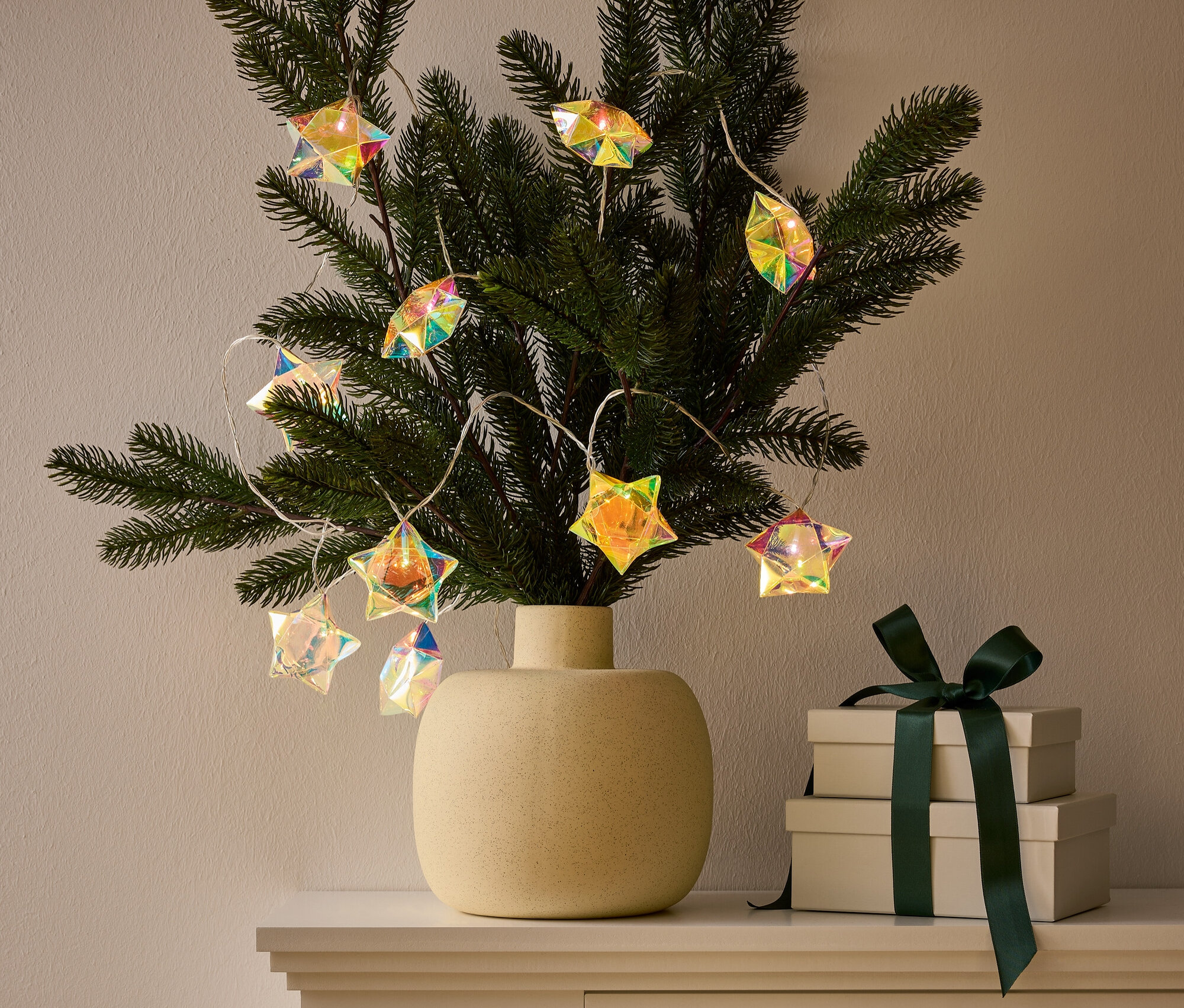 Yıldız şeklinde ışıklarla süslenmiş bir dalı olan bir vazo, koyu yeşil kurdeleli iki hediye kutusunun yanında bir dolabın üzerinde duruyor.