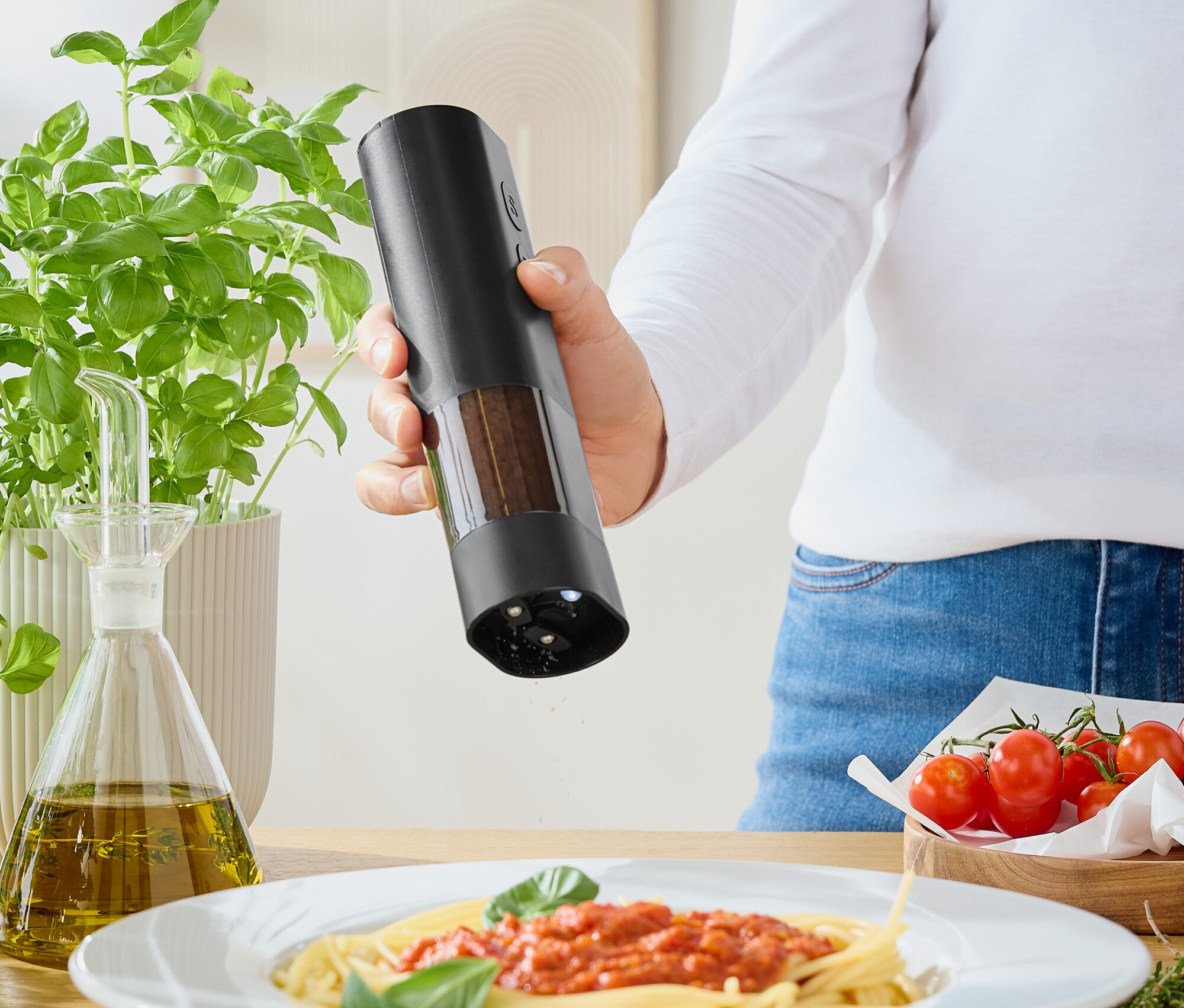 Kadın, değirmenden domates soslu spagettinin üzerine baharat serpiyor. Masanın üzerinde fesleğen, zeytinyağı ve domatesler var.