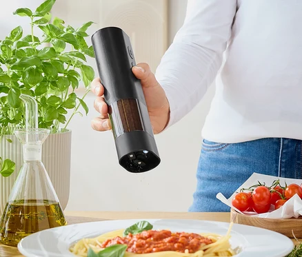 Kadın, değirmenden domates soslu spagettinin üzerine baharat serpiyor. Masanın üzerinde fesleğen, zeytinyağı ve domatesler var.