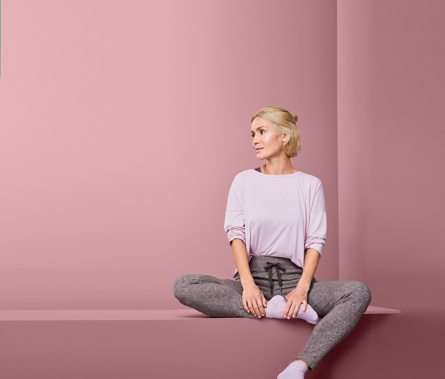 Sarışın bir kadın platformda bağdaş kurarak oturuyor ve rahat bir pantolon giyiyor.