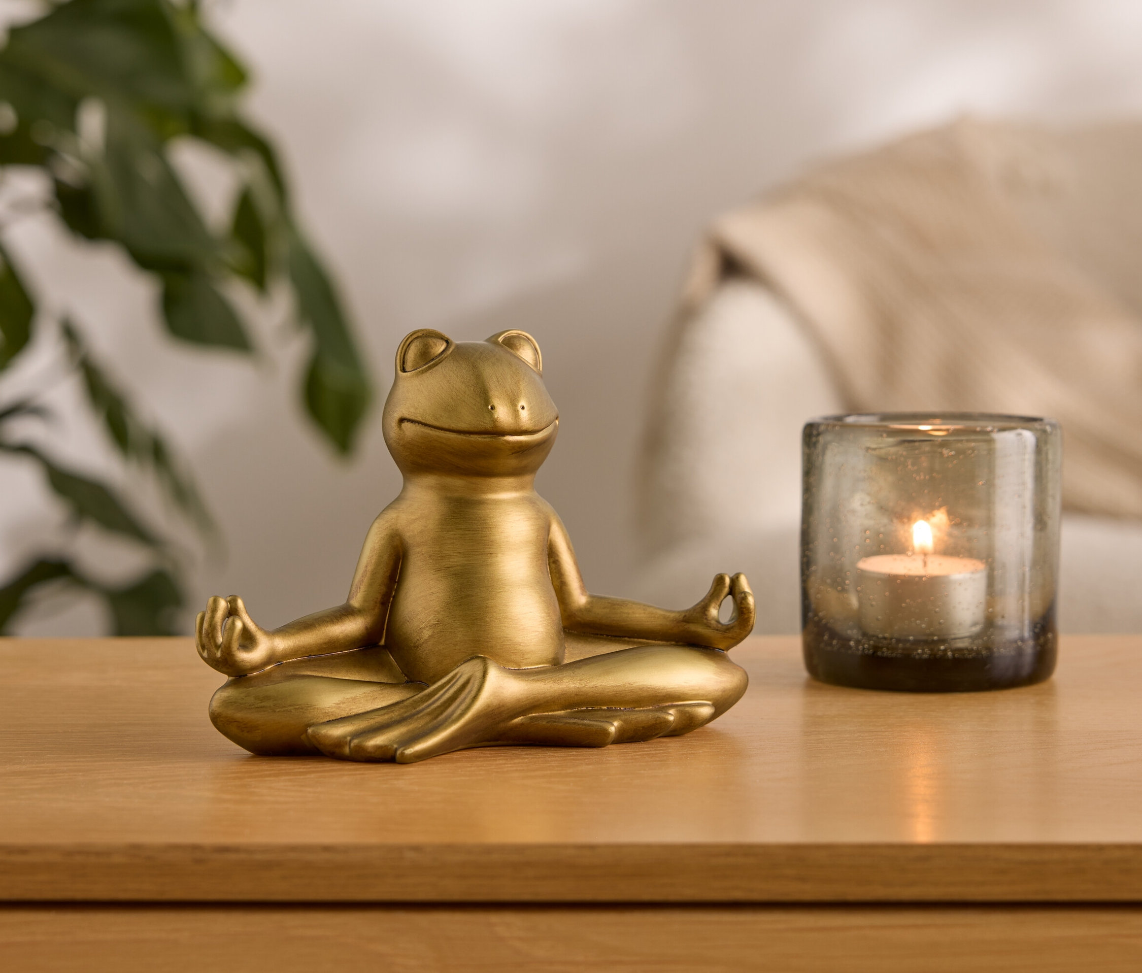 Altın renginde bir kurbağa heykeli, yanan bir mum bulunan cam bir kabın yanında meditasyon pozisyonunda oturuyor.