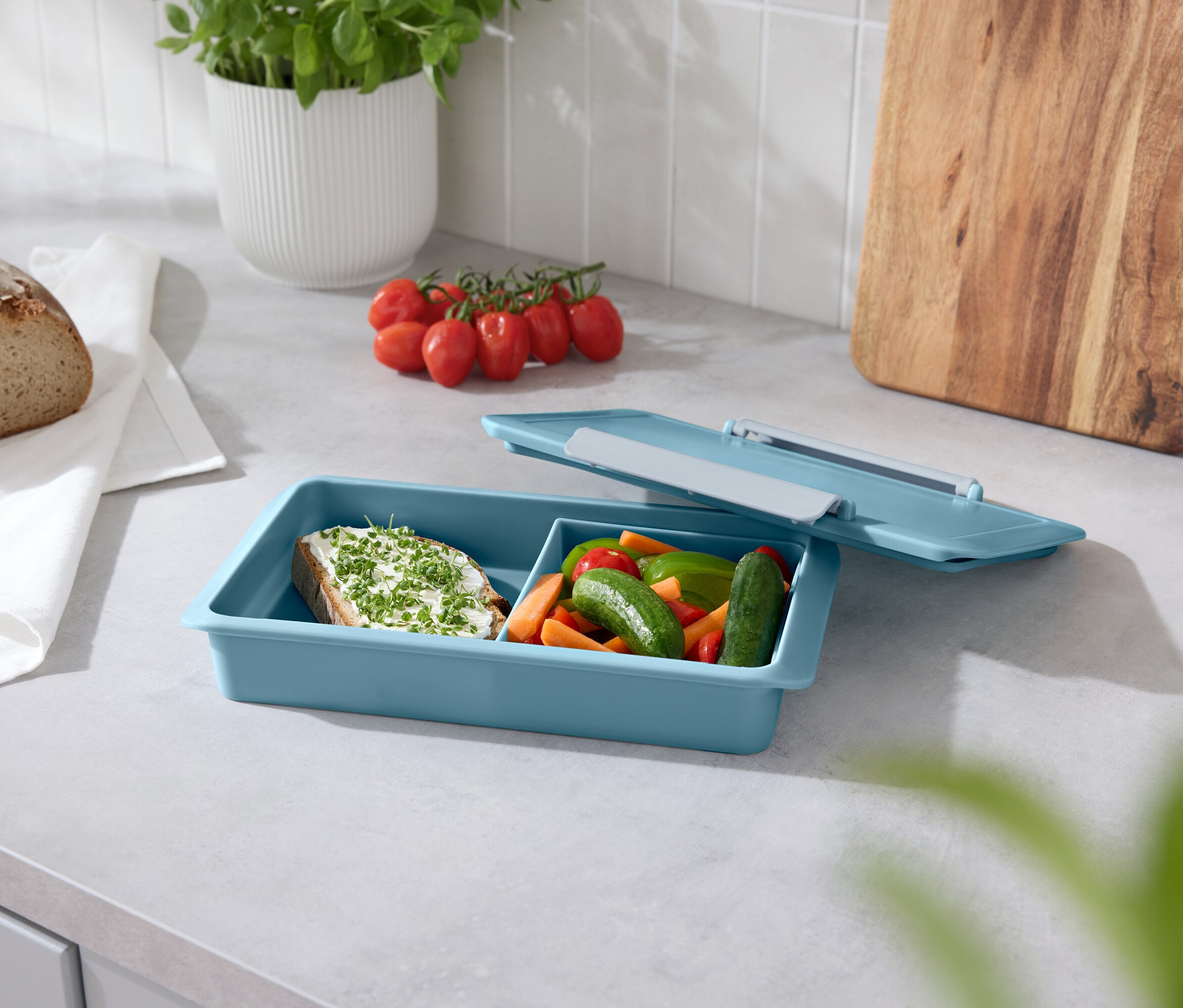 Ekmek, domates, fesleğen ve tezgah üzerinde açık kapaklı mavi bir atıştırma kutusu.