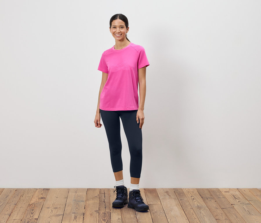 Kadın beyaz bir duvarın önünde duruyor, pembe bir tişört, koyu tayt ve yürüyüş ayakkabıları giyiyor.