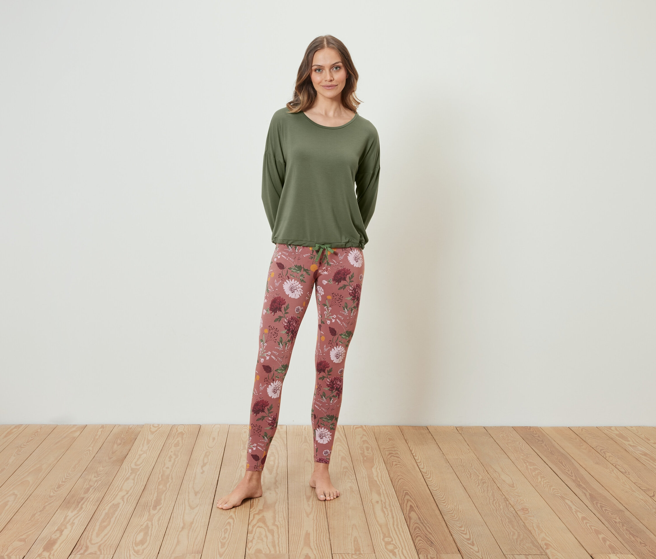 Ayakta duran bir kadın, üzerinde uzun kollu yeşil bir bluz ve çiçekli bir tayt var.