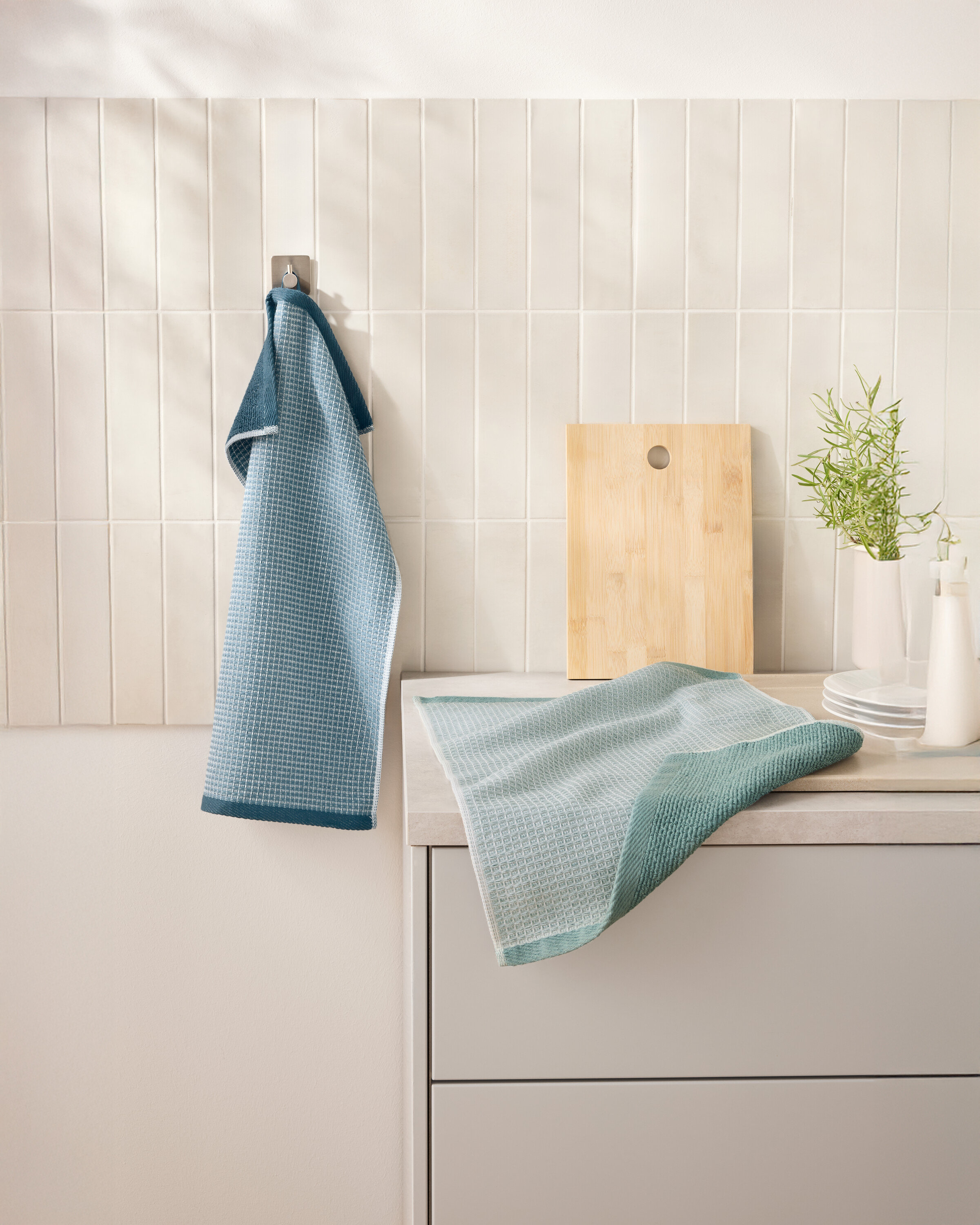 Mavi renkli 2 Waffelpiqué-Küchenhandtücher kesme tahtasının yanındaki bir kancada asılı duruyor. Başka bir mutfak havlusu mutfak tezgahında duruyor.