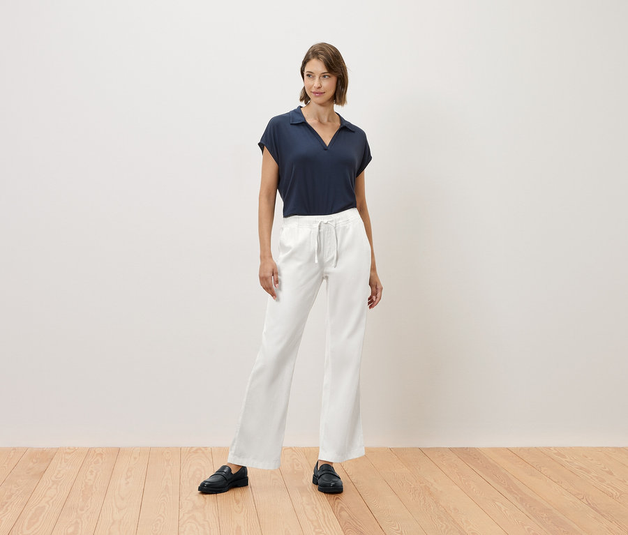 Bir kadın ahşap bir zeminde duruyor, lacivert bir tişört ve beyaz pantolon giyiyor.