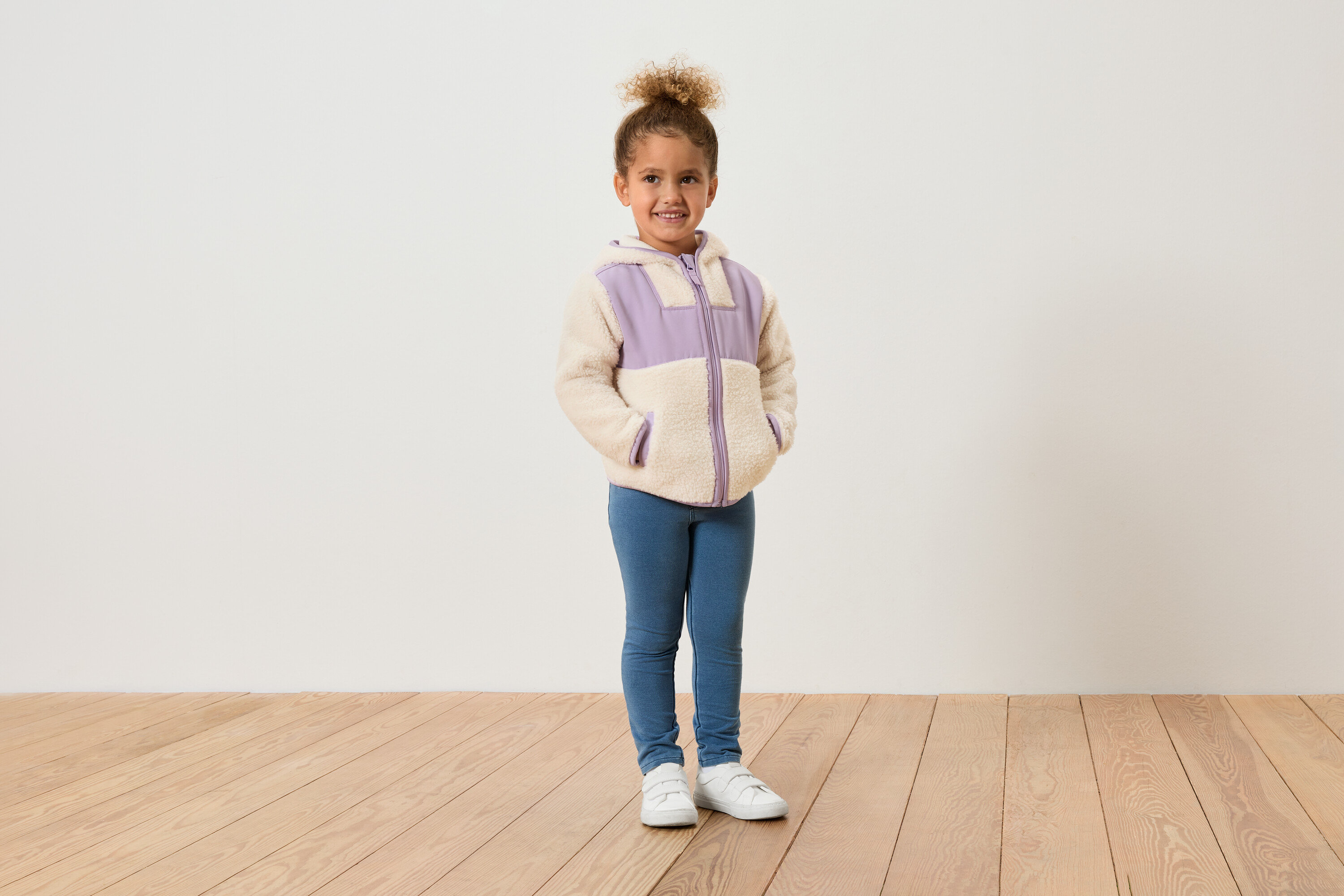 Genç bir kız beyaz bir duvarın önünde duruyor, lila detaylı bir polar ceket, mavi kot pantolon ve beyaz ayakkabılar giyiyor.