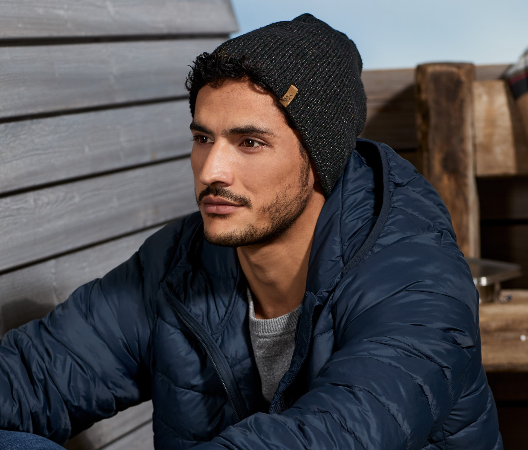 Adam ahşap zeminde oturuyor, üzerinde Termal Örgü Bere, Kırçıllı Antrasit, mavi bir ceket ve kot pantolon var.