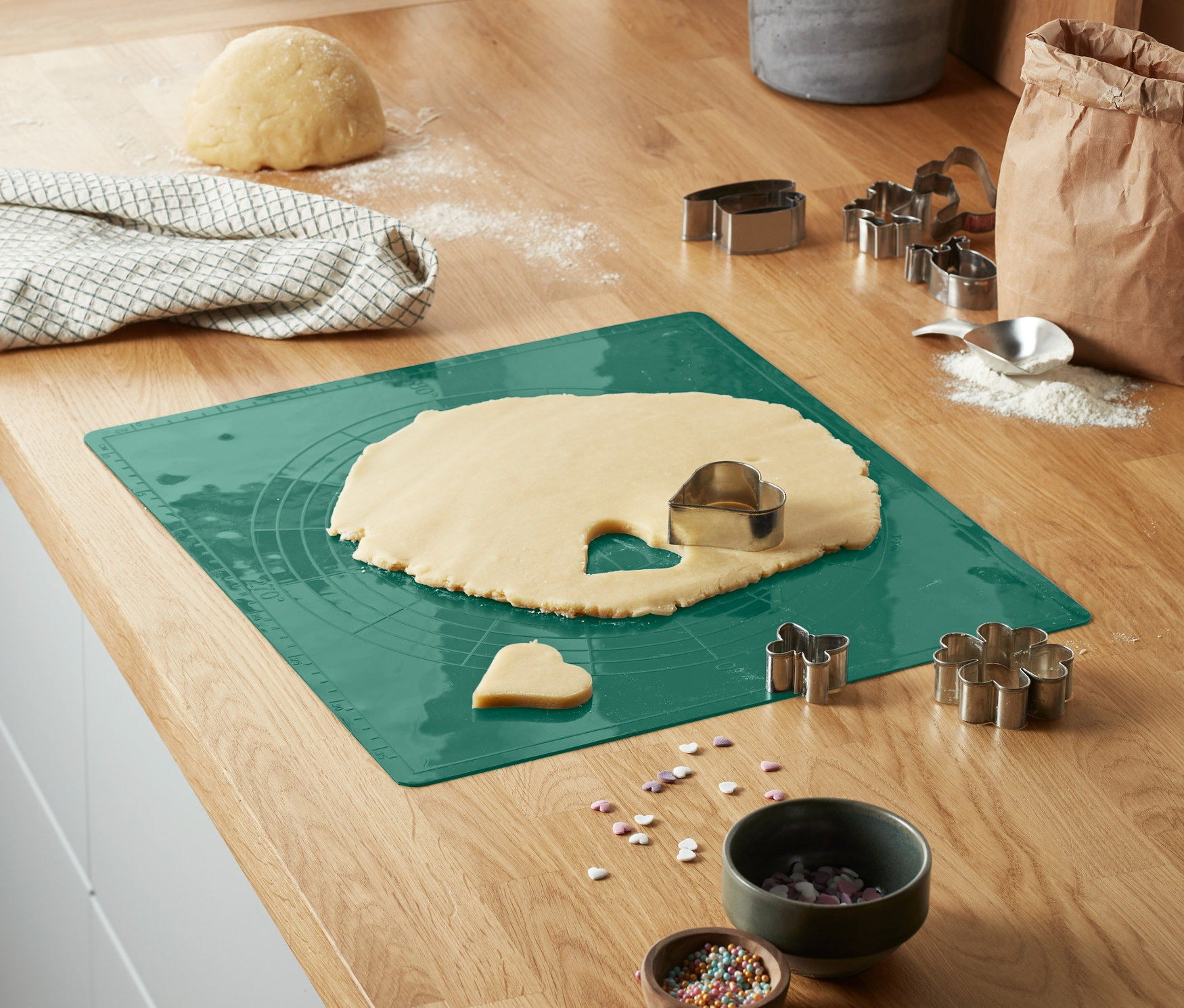 Mutfak tezgahında, açılmış hamur ve kurabiye kalıplarıyla birlikte yeşil bir Dayanıklı Pişirme Matı bulunmaktadır.