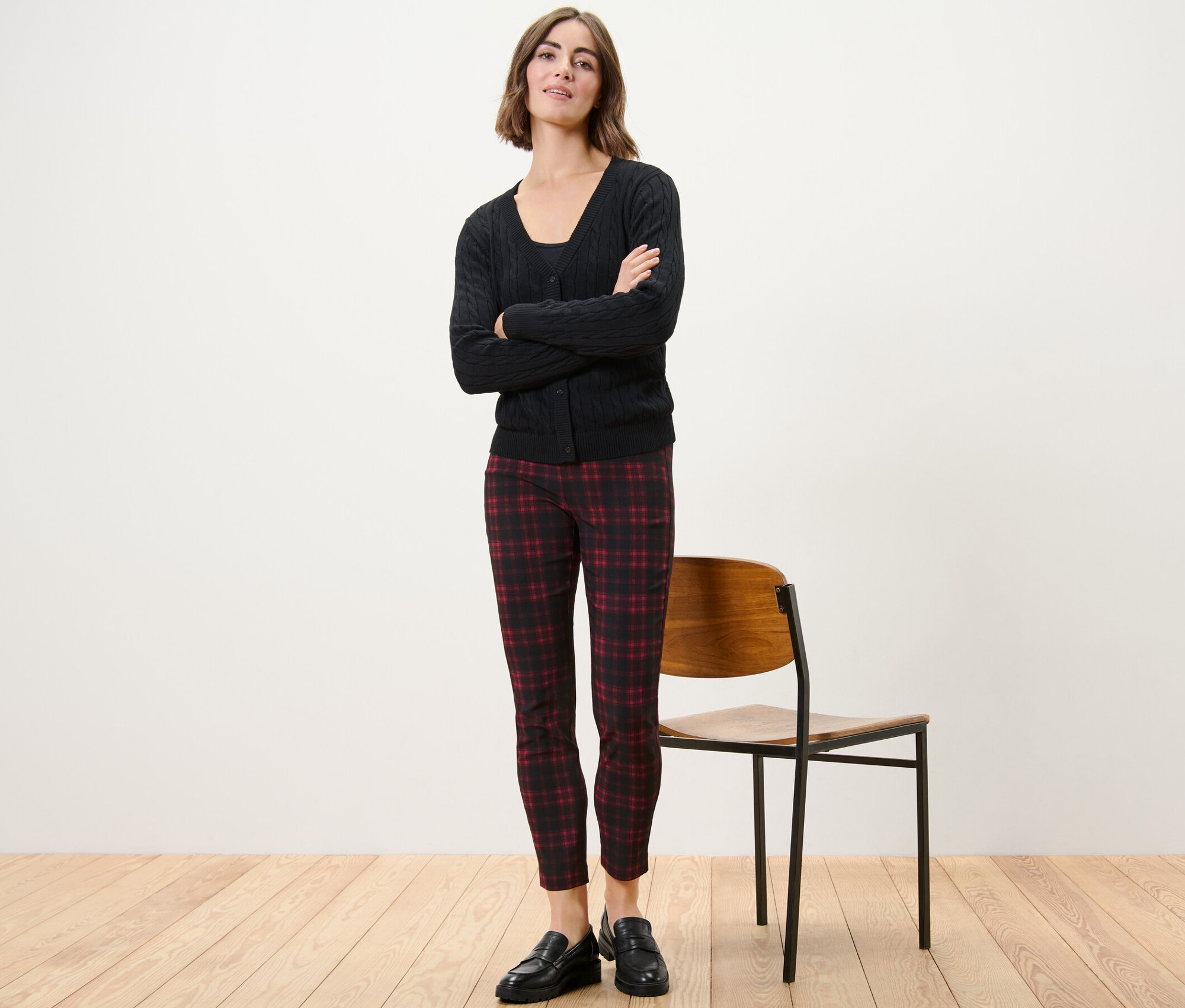 Bir kadın kolları çapraz bir şekilde duruyor, üzerinde kareli streç pantolon, siyah ceket ve siyah ayakkabılar var.