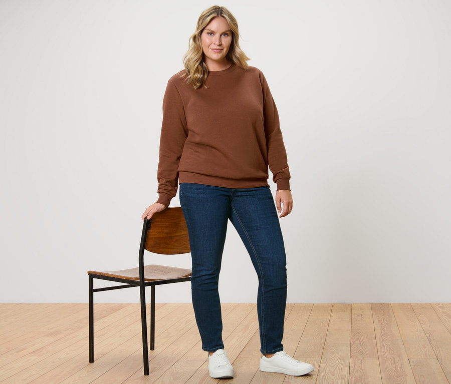 Bir kadın sandalyenin yanında duruyor, üzerinde kahverengi bir sweatshirt ve koyu mavi Slim Jeans – Fit »Emma« var.