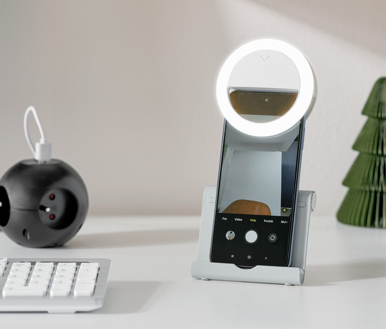 Gümüş renkli akıllı telefon ve tablet standı, beyaz bir masa üzerinde dairesel ışıklı bir akıllı telefonu tutuyor.