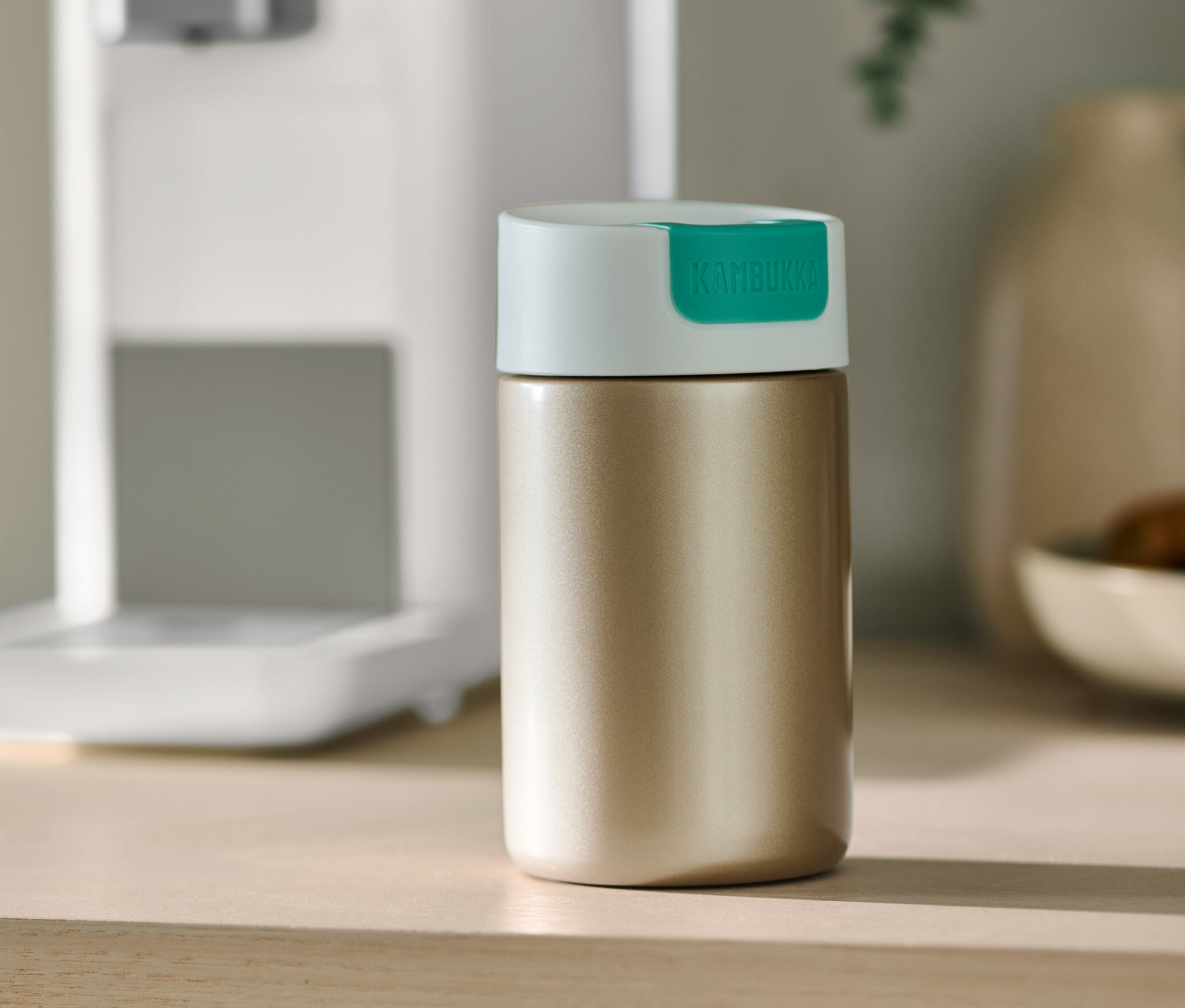 Altın rengi Kambukka To-Go bardak, bir masanın üzerinde kahve makinesinin yanında duruyor.