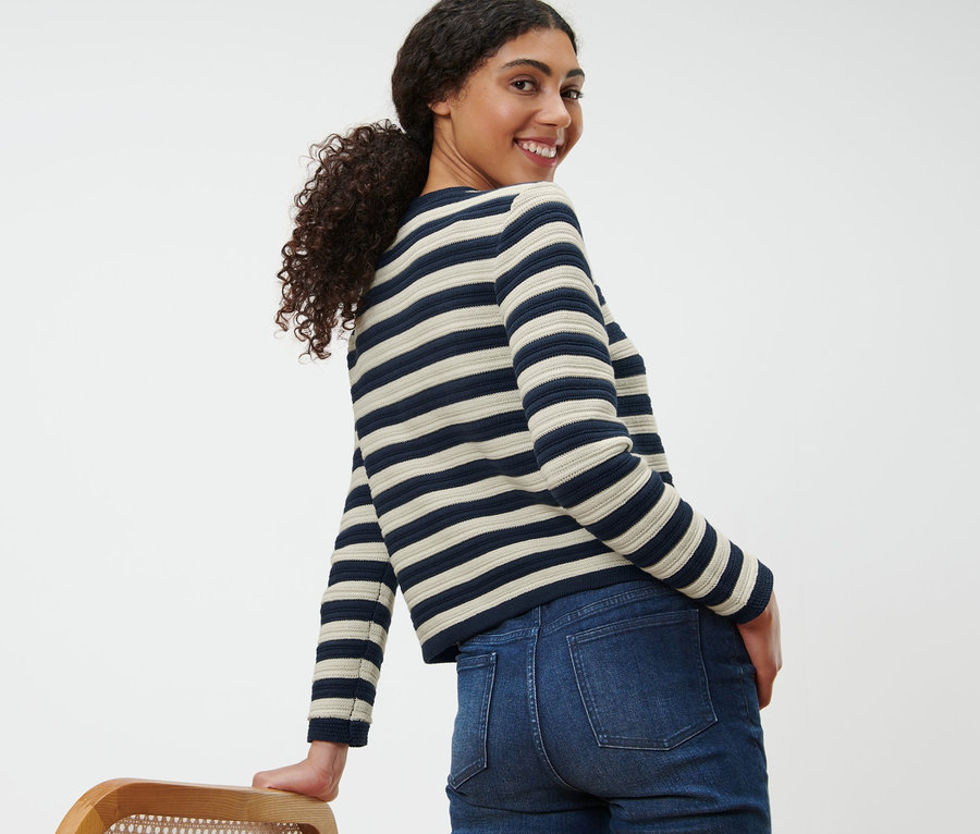 Gülümseyen bir kadın omzunun üzerinden bakıyor, dokulu örgü hırka ve kot pantolon giyiyor.