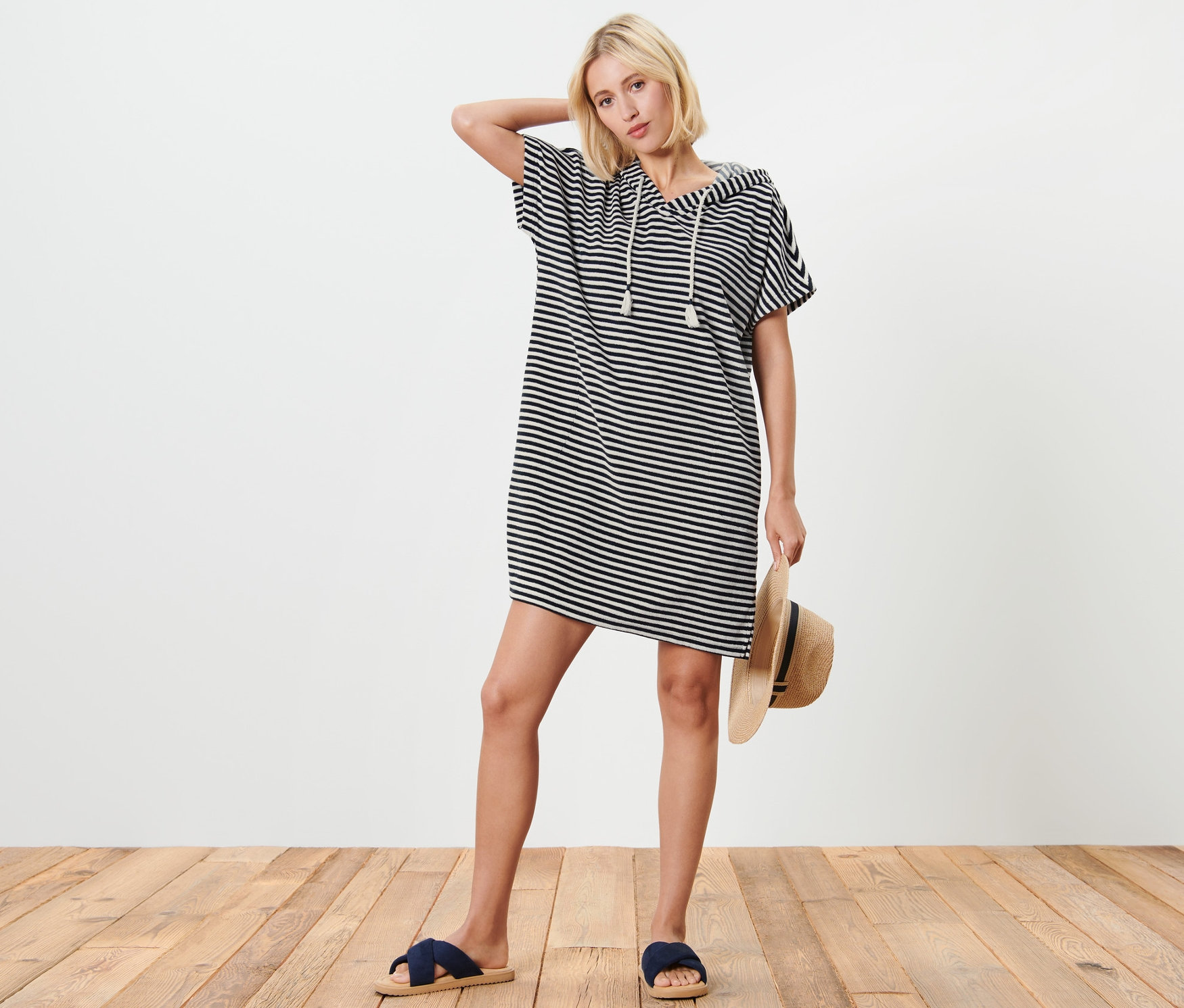 Sarışın bir kadın, mavi-beyaz çizgili, kapüşonlu havlu kumaş plaj elbisesiyle poz veriyor, bir hasır şapka tutuyor ve mavi terlikler giyiyor.